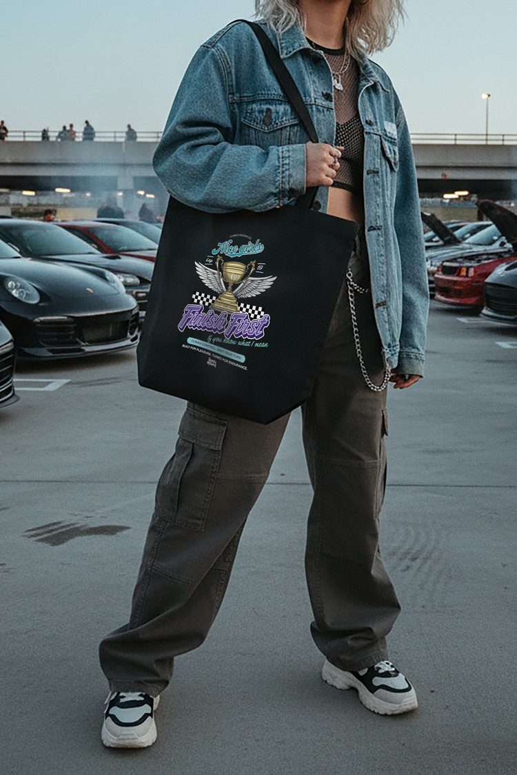 Woman holding a black tote bag with a car design in a parking lot with cars and a bridge in the background.